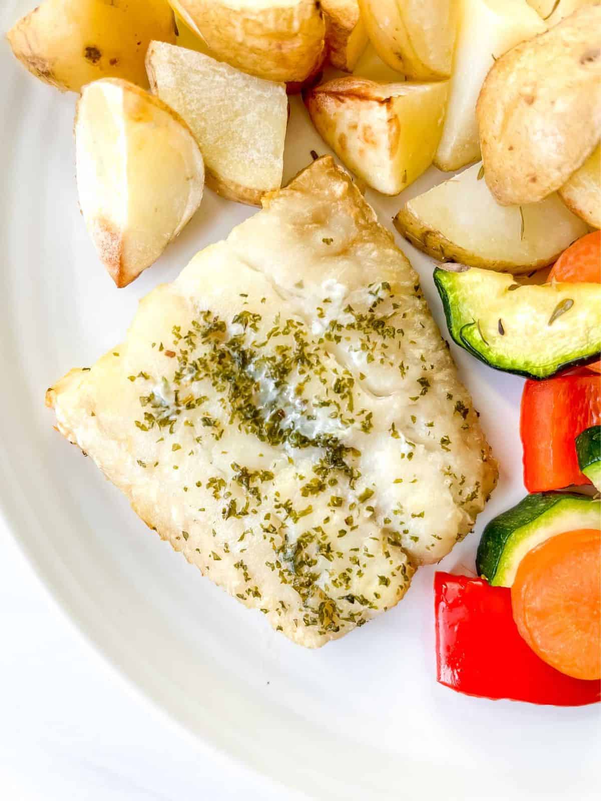 cod fillet on a white plate with potatoes and roasted vegetables.